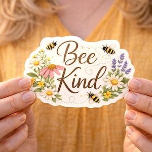 May include: A white sticker with the words "Bee Kind" in brown script, surrounded by illustrations of bees, daisies, lavender, and other flowers. The sticker has a floral border and is held up by a person.