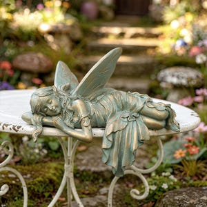 Könnte beinhalten: Eine bronzefarbene Feenstatue mit detaillierten Flügeln und fließendem Kleid, die auf einem weißen, runden Tisch ruht. Die Fee liegt mit geschlossenen Augen. Der Tisch hat verzierte, geschwungene Beine, im Garten.