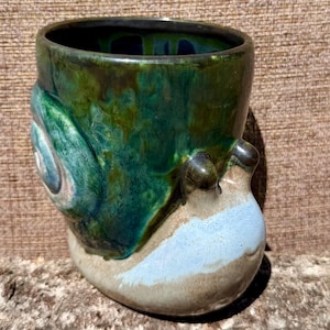 May include: A ceramic mug shaped like a snail with a glossy green and blue glazed shell. The base of the mug is a light beige color with a smooth, rounded form, featuring two small protrusions at the top representing the snail's eye stalks.