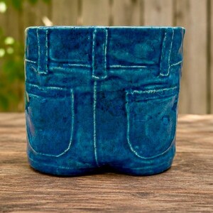 May include: A deep blue ceramic planter shaped like the back of a pair of denim jeans, featuring molded details such as two back pockets, a waistband, and belt loops with a glossy, textured finish that mimics the appearance of fabric seams.