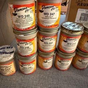 May include: A collection of Duncan E-Z Flow ceramic glaze jars in 4 ounce sizes. The labels feature various woodtone colors including Country Pine WD 246, Walnut WD 247, Fruitwood WD 248, and WD 253. Each jar has a metal lid and orange branding.