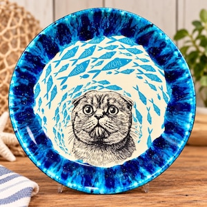 May include: A round ceramic plate featuring a central illustration of a Scottish Fold cat face surrounded by a school of small blue fish. The plate has a wide, vibrant blue rim with a marbled, watercolor-like texture and a cream-colored center.