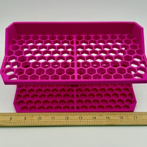 Puede incluir: Un organizador magenta brillante, impreso en 3D, con un patrón de panal. El organizador tiene una bandeja en la parte superior y una base, ambas con el mismo diseño hexagonal. Una regla de madera en la parte inferior muestra medidas en centímetros.