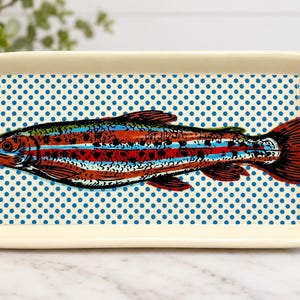 May include: A rectangular cream-colored ceramic serving tray with integrated side handles. The center features a detailed illustration of a rainbow trout with red, blue, and black markings, set against a background of small blue polka dots.