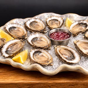 May include: A large, scalloped, light-colored ceramic platter filled with crushed ice, holding eleven raw oysters in their shells, three lemon wedges, and a small metal ramekin containing a red onion mignonette sauce.