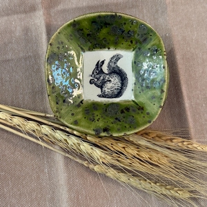 May include: A small, square ceramic dish with a green speckled glaze. The center features a white square with a black and white illustration of a squirrel. The dish is surrounded by wheat stalks.