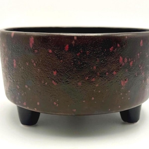 May include: A brown ceramic bowl with a speckled red pattern and three black legs. The bowl has a textured surface and a dark interior. This decorative bowl is suitable for plants or as a decorative accent.