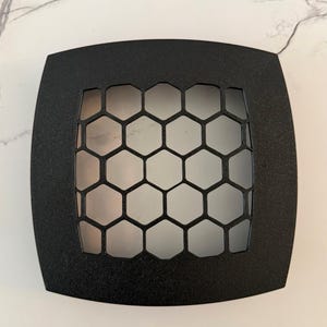 May include: A black, square-shaped metal vent cover with rounded corners. The center features a honeycomb pattern. The vent cover is likely used for home decor or ventilation purposes.