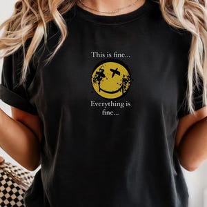 Pode incluir: Camiseta preta com o texto "This is fine... Everything is fine..." e um gráfico de carinha sorridente amarela. O gráfico tem olhos cruzados pretos e um design desgastado. A palavra "BLACK" está impressa no canto superior esquerdo.