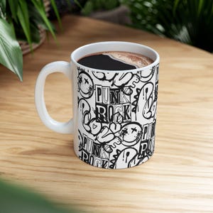 Pode incluir: Uma caneca de cerâmica branca com uma alça preta e o texto "But First, Coffee" em negrito preto. A caneca está em um fundo desfocado. A caneca tem aproximadamente 10 cm de altura.