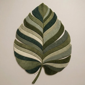 May include: A large, leaf-shaped rug in shades of green, olive, and cream. The rug is made of felt and features a layered design, with each section a different shade. The stem of the leaf is a darker green.