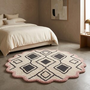 May include: A round rug with a scalloped edge in a bedroom setting. The rug features a cream-colored center with gray diamond shapes and a pink border. The bed has cream-colored bedding.