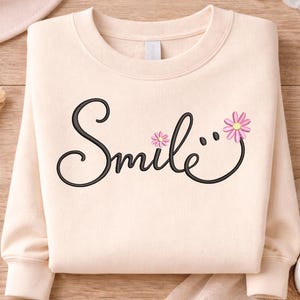Może przedstawiać: Kremowa bluza z napisem "Smile" w czarnym skrypcie, zaakcentowana dwoma różowymi motywami kwiatowymi. Bluza jest starannie złożona na drewnianej powierzchni, co sugeruje swobodny, wygodny styl.