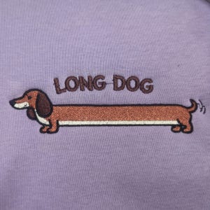 May include: A lilac-colored shirt featuring an embroidered dachshund dog with a long body. The text "LONG DOG" is embroidered above the dog. The dog is brown with a white underbelly and black accents.