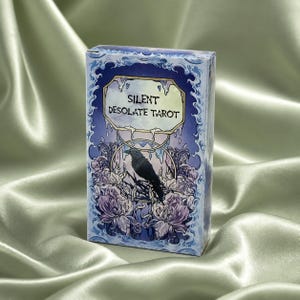 Puede incluir: Una caja de cartas del tarot con un diseño floral azul y morado. La caja presenta el texto "SILENT DESOLATE TAROT" y una ilustración de un cuervo negro posado en una rama. El fondo es una tela de satén.