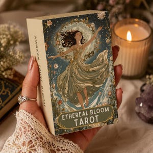 Puede incluir: Una caja de cartas de tarot beige con las palabras "Ethereal Bloom Tarot" en la parte delantera. La caja presenta una ilustración de una mujer con un vestido fluido con una corona floral, sobre un fondo de cielo nocturno. Las palabras "Awaken, Trust, Bloom" están impresas en el lateral.