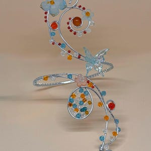 May include: A silver-toned arm cuff with a floral and butterfly design. The cuff features light blue flowers, orange and red beads, and a translucent blue butterfly. The design incorporates a spiral and an oval shape, with a large orange bead at the base.