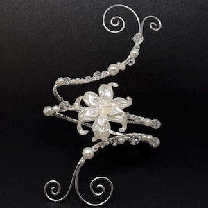 May include: A silver arm cuff with a central white flower design. The cuff is embellished with faux pearls and clear crystals, with decorative swirls at each end. The piece is set against a black background, highlighting its delicate details.