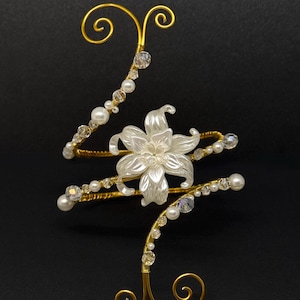 May include: A gold-colored arm cuff with a white flower and pearl accents. The cuff features a floral design with a central flower and is adorned with pearls and clear crystals. The design has a spiral shape with decorative swirls.