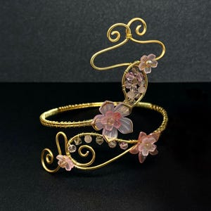 May include: A gold-toned arm cuff with a floral design. The cuff features delicate pink flowers and clear beads. The design includes swirling wire accents and is set against a dark background.