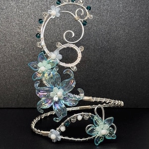 May include: A silver wire ear cuff featuring iridescent blue and clear glass flower petals, small white beads, and dark teal crystal accents. The design includes swirling wire patterns and a coiled structure that wraps around the ear shape.