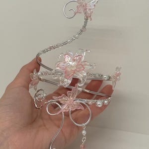 Pode incluir: Um adorno de cabeça decorativo prateado com delicadas flores e borboletas cor-de-rosa. O design incorpora pétalas transparentes iridescentes, pérolas falsas e uma corrente pendurada de contas. O adorno de cabeça é segurado em uma mão contra um fundo liso.