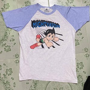 May include: A white and light blue raglan t-shirt featuring the cartoon character Mighty Atom. The graphic shows the character in a dynamic pose with the text "MIGHTYATOM" above him. The shirt is made of a soft material.