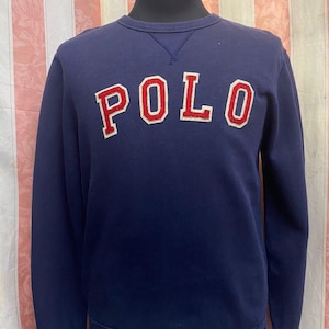 May include: Navy blue sweatshirt with the word "POLO" in large, red, outlined letters across the chest. The sweatshirt has a crew neck and ribbed cuffs and hem. The number "67" is visible on the lower left cuff.