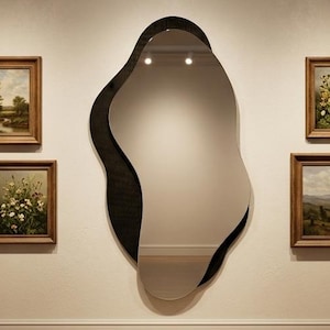 Op de afbeelding: Interieur van een kunstgalerie met een grote, onregelmatig gevormde spiegel met een zwarte rand. Verschillende ingelijste landschapsschilderijen worden op de witte muren getoond. Betonnen banken staan op de houten vloer, verlicht door rails.