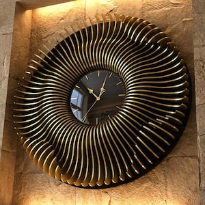 Può includere: Un grande orologio da parete rotondo con quadrante nero e lancette dorate. L'orologio è circondato da un design decorativo a spirale di strisce metalliche dorate. È montato su un muro di pietra strutturata marrone chiaro.