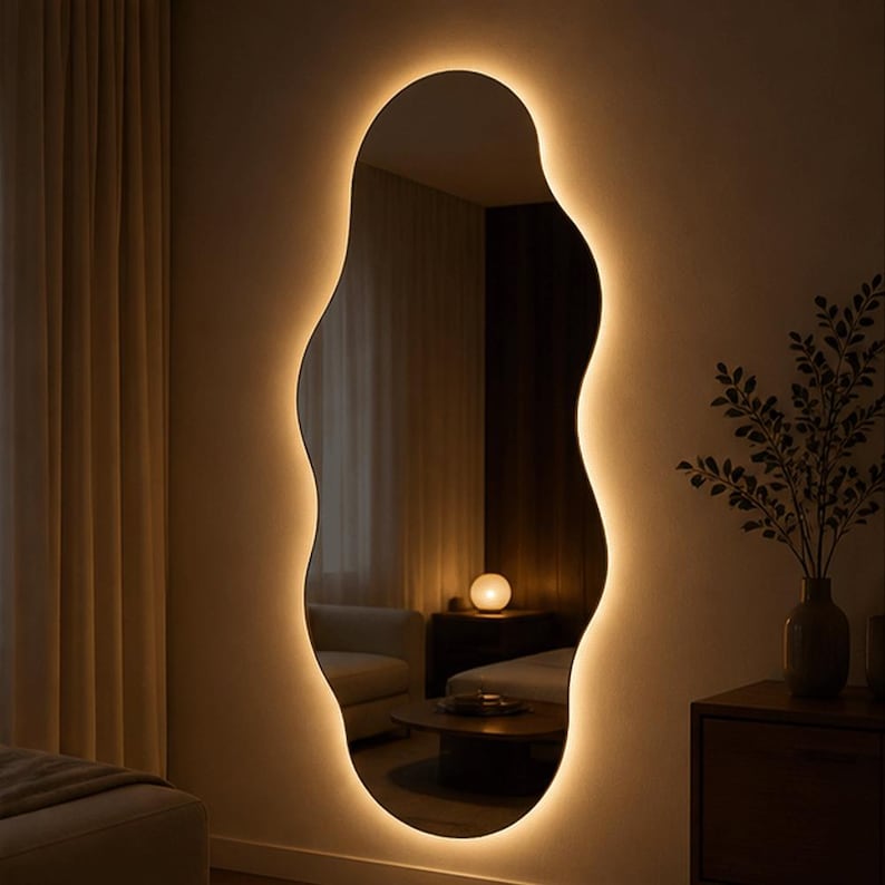May include: A large, organically shaped mirror with a warm, glowing LED light border. The mirror is mounted on a neutral-colored wall, reflecting a room with a sofa, table, and decorative items. The overall aesthetic is modern and minimalist.