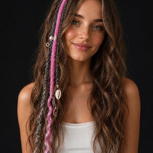 Peut inclure: Une femme aux longs cheveux bruns ondulés avec des tresses roses et argentées. Les tresses sont ornées de breloques décoratives, dont un coquillage et des perles. Elle porte un débardeur blanc sur fond noir.