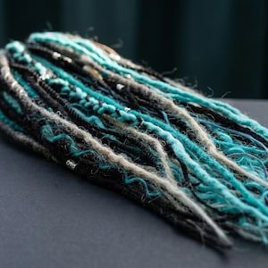 Könnte beinhalten: Ein Set aus synthetischen Dreadlocks mit einer Mischung aus schwarzen, türkisfarbenen und hellgrauen Strähnen. Die texturierten Haarverlängerungen enthalten geflochtene Abschnitte und sind mit kleinen silberfarbenen Metallperlen verziert.