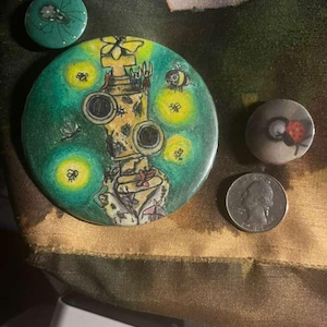 May include: A collection of handmade art buttons. The largest button features a whimsical illustration of a machine with bees and glowing lights on a green background. Other buttons include a spider and a ladybug.