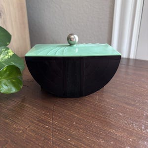 May include: A rectangular box with a black base and a light green lid. The lid features a silver spherical knob and curved, radiating lines. The black base has a central vertical ribbed panel flanked by two semi-circular, stepped-in sections.