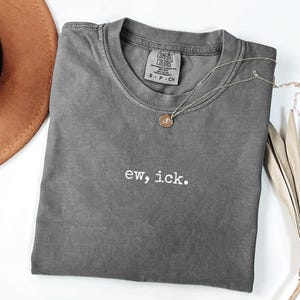 Puede incluir: Una camiseta gris Comfort Colors con la frase "ew, ick." en letras blancas. Un collar dorado con un pequeño colgante descansa sobre la camiseta. Un sombrero marrón y una planta seca están en el fondo.
