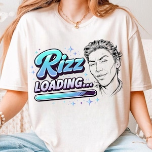 May include: Cream-colored t-shirt featuring the text "Rizz LOADING..." in a blue and teal gradient font, with a stylized illustration of a man's face on the right. The design includes stars and a loading bar.