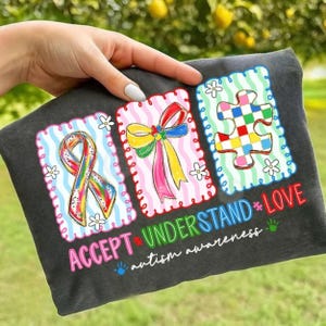 May include: Dark gray t-shirt with three colorful graphic designs. The designs include a rainbow ribbon, a bow, and a puzzle piece, all framed with decorative borders. The words "ACCEPT UNDERSTAND LOVE" and "autism awareness" are printed on the shirt.