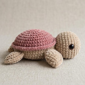 Könnte beinhalten: Eine handgefertigte Schildkröte aus Plüsch in Rosa- und Beigetönen. Die Schildkröte hat einen strukturierten Panzer, einen runden Kopf mit einem einzelnen schwarzen Auge und vier flossenartigen Anhängseln. Das Kuscheltier ist weich und kuschelig.
