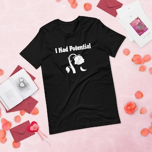 May include: Black t-shirt with the text "I Had Potential" above a white illustration of a wilting rose. The shirt is a short-sleeved crew neck style. The image also includes red envelopes, rose petals, and cards.