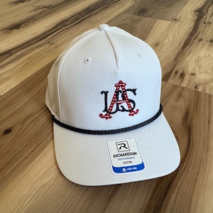 Pode incluir: Boné de beisebol branco com aba reta e detalhe de corda preta. O boné apresenta as letras bordadas "IAS" em vermelho e preto. Uma pequena etiqueta com a marca "Richardson" está anexada.