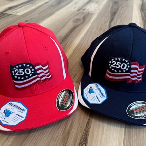 Puede incluir: Dos gorras de béisbol, una roja y otra azul marino, con detalles blancos. Cada gorra presenta un parche con el número "250" y un diseño de bandera estadounidense. Las gorras tienen una visera curva y un ajuste estructurado.