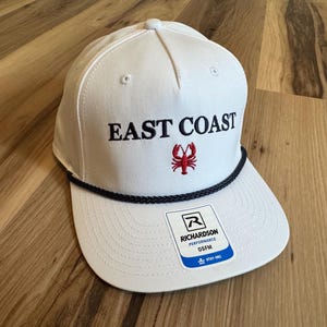 Op de afbeelding: Witte baseballcap met de tekst "EAST COAST" in zwart en een rode kreeftafbeelding. De cap heeft een zwart touwdetail langs de rand en een Richardson Performance-label.