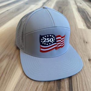 Puede incluir: Gorra de béisbol gris claro con visera plana. La gorra presenta un parche con un diseño estilizado de la bandera estadounidense y el número "250" en blanco. La gorra tiene pequeños orificios de ventilación.