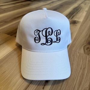 May include: A white baseball cap with a curved peak. The cap displays a navy blue monogram in a decorative, flowing script. The cap is made of a smooth material and has a button on top.