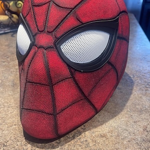 May include: A red and black Spider-Man mask with white mesh eye lenses. The mask has a textured, web-like pattern and black outlines. The mask is a cosplay or costume accessory.