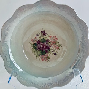 May include: A vintage, iridescent, scalloped-edge bowl. The bowl features a floral design of purple and pink violets in the center. The rim has a subtle, embossed pattern. The bowl is displayed on a clear stand.