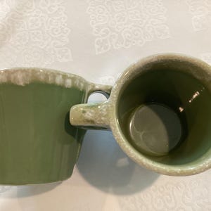 May include: Two green ceramic mugs with a light-colored rim and handle. The mugs are angled towards each other, showcasing their interior and exterior design. The mugs are on a white surface.