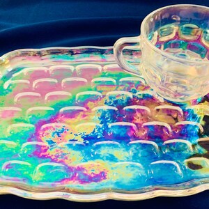 May include: Iridescent glass teacup and tray set. The tray has a rectangular shape with a scalloped edge and a raised pattern. The teacup has a handle and a matching iridescent finish, reflecting colors like blue, pink, and yellow.