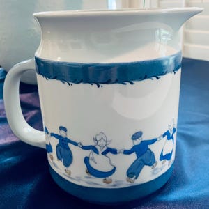 May include: A white ceramic pitcher featuring a blue painted band at the top and bottom. The center of the pitcher displays a repeating pattern of figures in traditional blue and white clothing holding hands while dancing on a light blue surface.